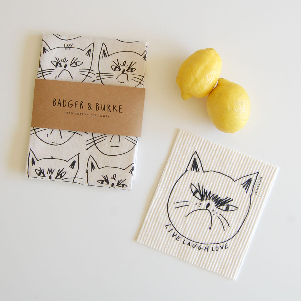 a product photography shot featuring a tea towel with Snitty Kitty's face, and a swedish sponge cloth with Snitty Kitty's face and the text "Live Laugh Love"