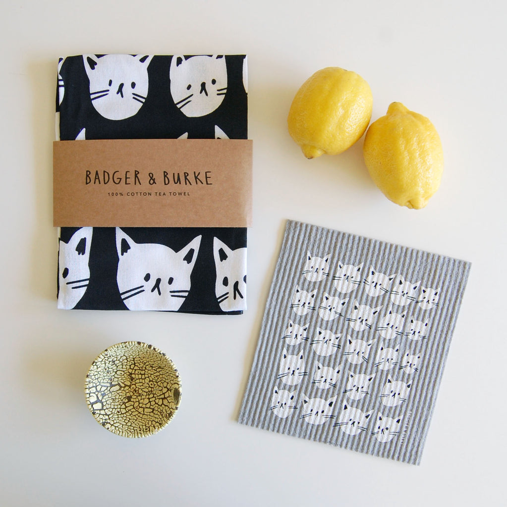 a product photo shot of a black tea towel with white cat faces, and a grey swedish sponge cloth with the same pattern of white cat faces. Surrounding the products are two lemons and a small pinch bowl with a yellow finish. 