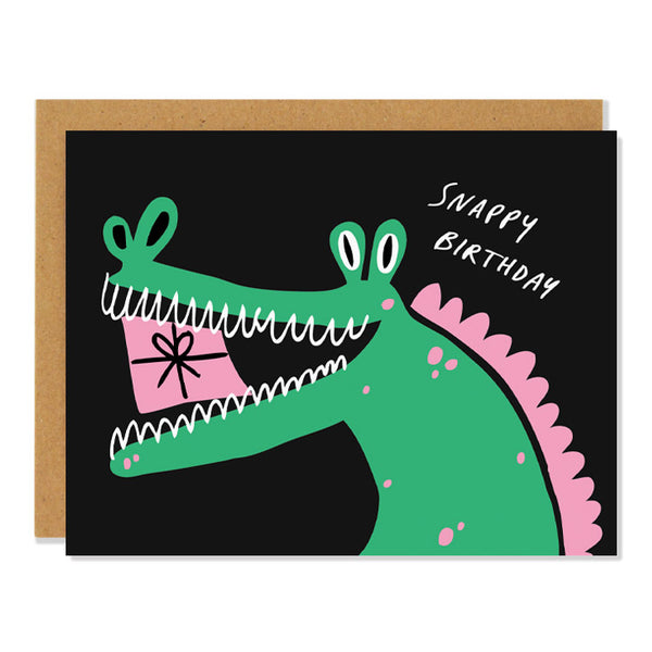 a birthday greeting card featuring an illustration of a cheery alligator in green and pink with a present in it's mouth. text in the top right corner reads: "Snappy Birthday"