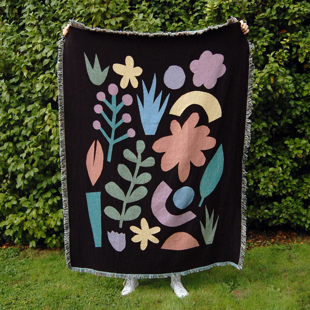 A black woven blanket with colorful abstract floral and botanical designs in front of a green hedge, with two hands holding the corners at the top.