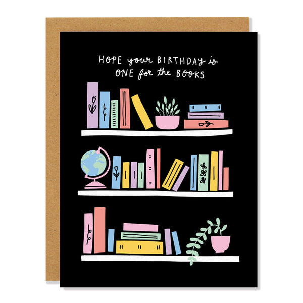 a birthday greeting card featuring an illustration of bookshelves with pastel coloured books, houseplants, a globe. Text above reads "hope your birthday is one for the books"