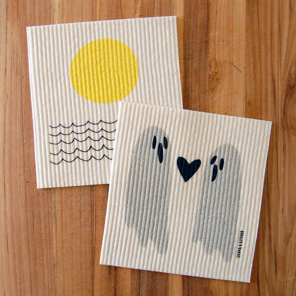 two swedish sponge cloths on wood background, left: a yellow sun above black lines representing waves of the ocean, right: two grey ghosts with a black heart inbetween