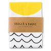 a tea towel featuring minimalist illustrations of a yellow sun and black wavy lines representing the sea, on white cotton.