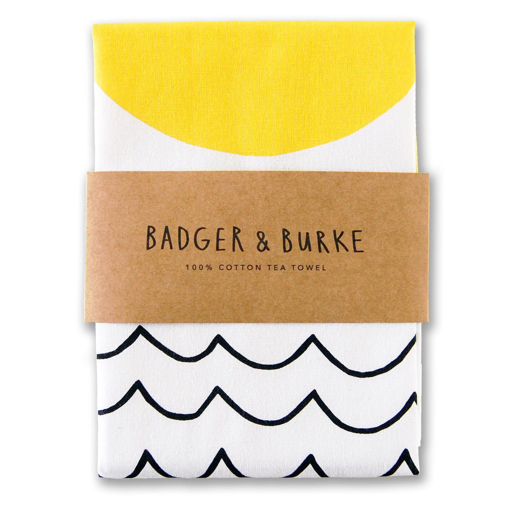 a tea towel featuring minimalist illustrations of a yellow sun and black wavy lines representing the sea, on white cotton.