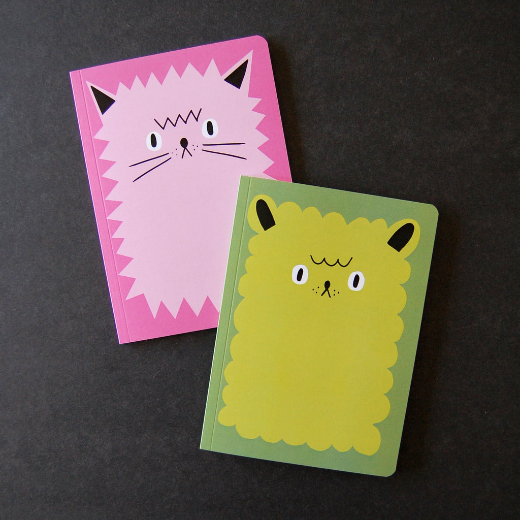 Two cute illustrated notebooks with one cat face and one dog face, one pink with a light pink spiky cat and a dog is green with a yellow furry cat, placed on a black background.