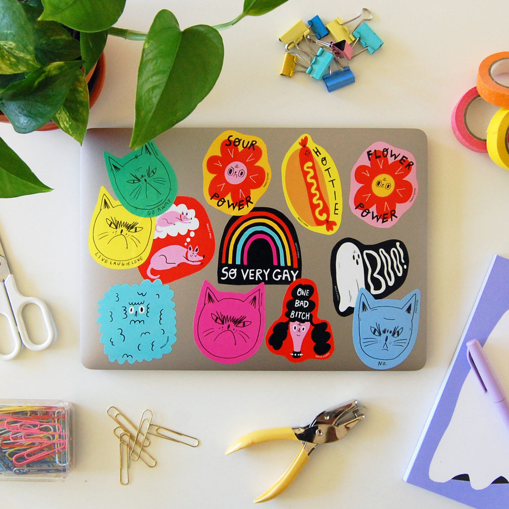 Colorful stickers on a laptop with various designs, including cats, a hot dog, flowers,, rainbows, and ghosts, surrounded by office supplies like scissors, paper clips, and a plant.