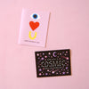 Two Valentine's Day cards on a pink background. The top card is light pink with a graphic of an eye, heart, and a U with the words "HAPPY VALENTINE'S DAY." The bottom card is black with a cosmic design and the words "OUR LOVE IS COSMIC, HAPPY VALENTINE'S DAY."