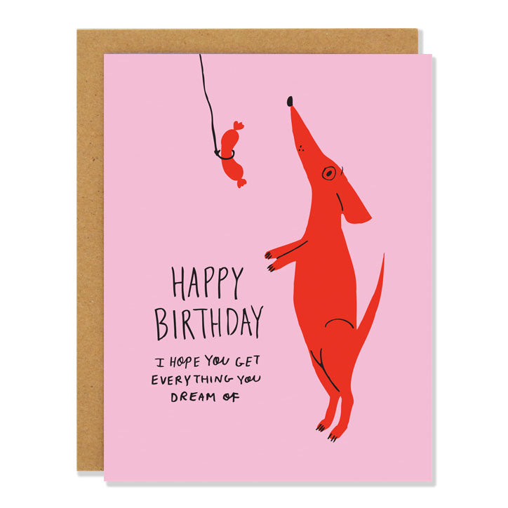 A birthday card with a pink background and a red dog standing on two legs looking up at a sausage on a string with the text "Happy Birthday I hope you get everything you dream of"