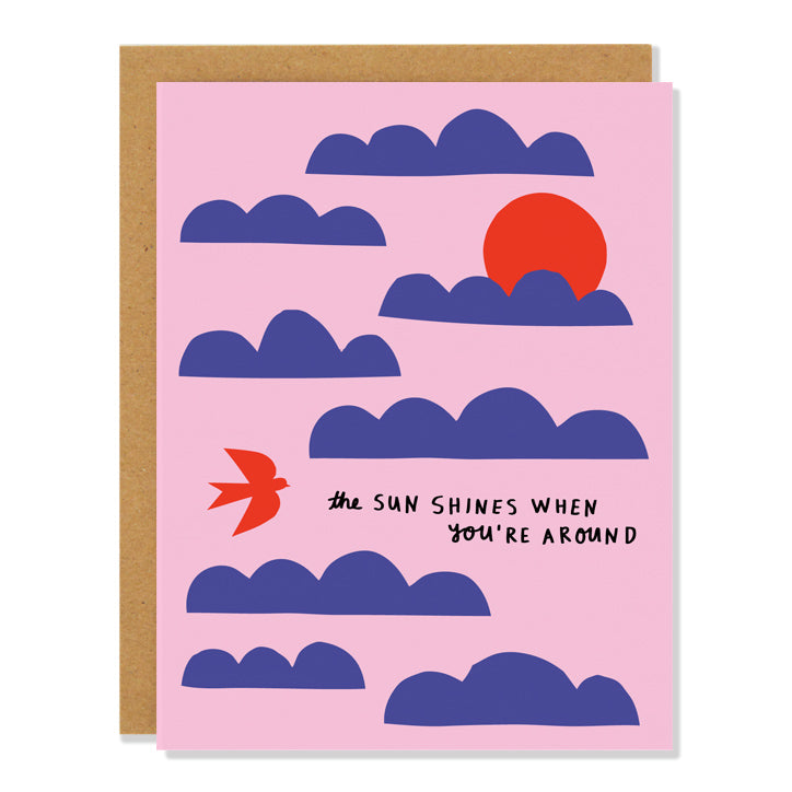 a love and friendship greeting card featuring minimalist illustrations of dark blue clouds and red skies and a red bird on a light pink background. The handwritten text reads: "the sun shines when you're around". 